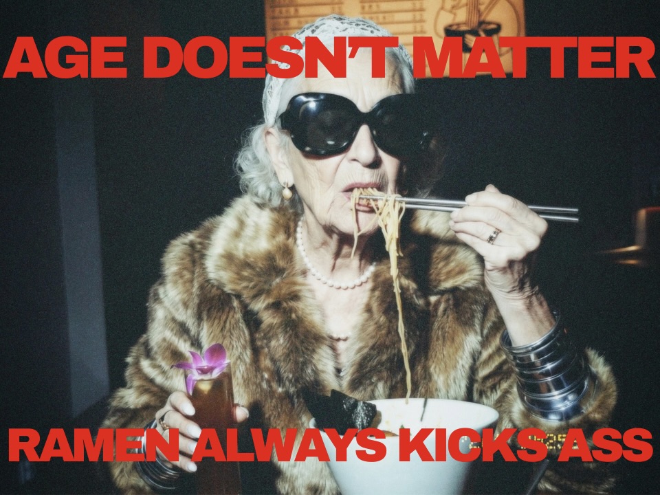 Starsza kobieta w ciemnych okularach i futrze je ramen pałeczkami; czerwony napis: AGE DOESN’T MATTER / RAMEN ALWAYS KICKS ASS